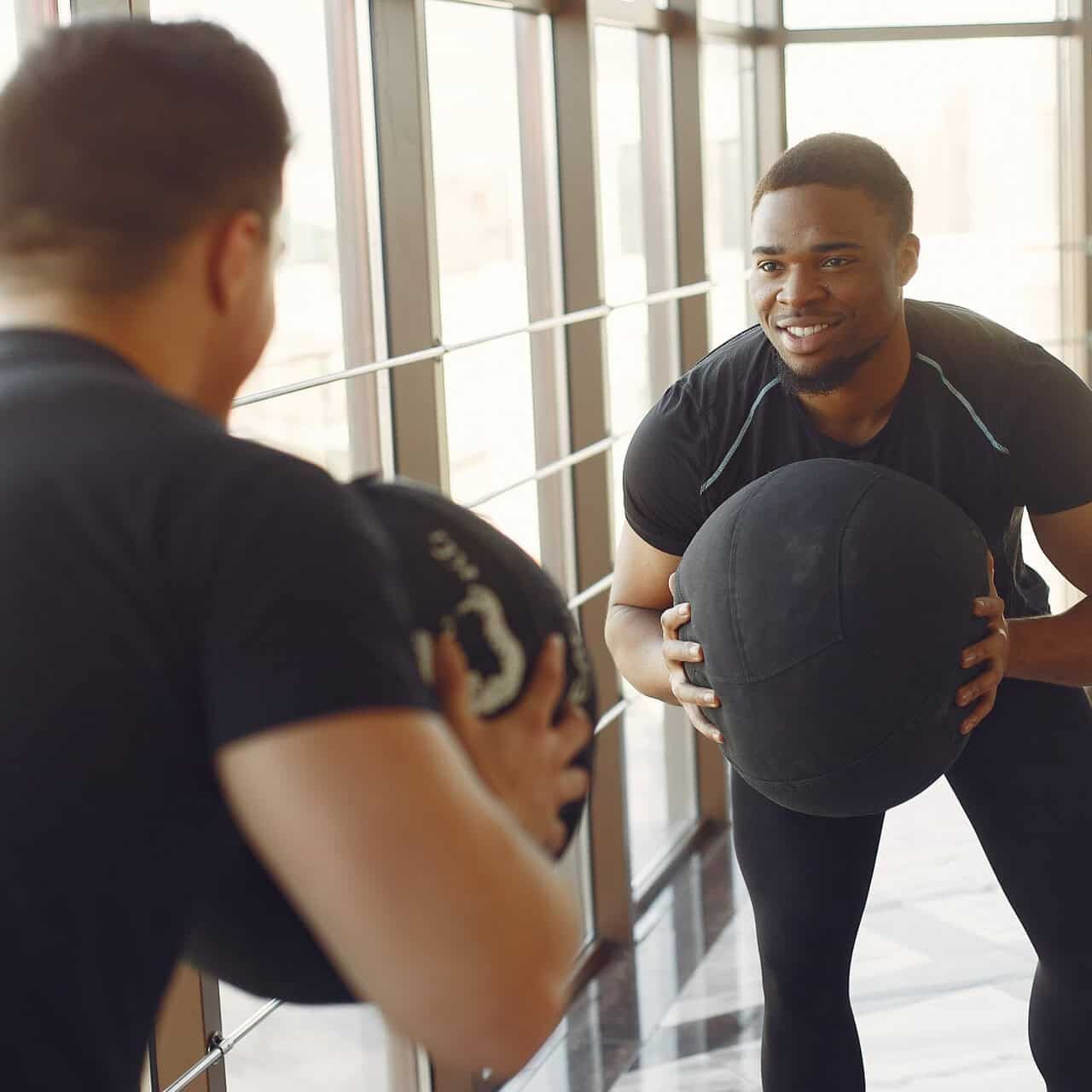 Líder de treino a ajudar a pessoa a realizar exercício com bola medicinal no ginásio, promovendo saúde, fitness e bem-estar.